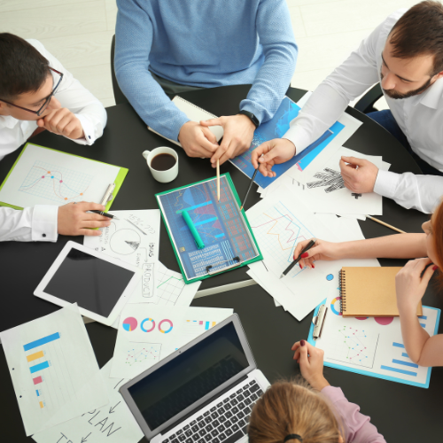 A group of five professionals sitting around a circular black table, collaborating on a project. The table is covered with charts, graphs, tablets, laptops, notebooks, and a cup of coffee. Each person is engaged in discussion or pointing to data visualizations, suggesting teamwork and brainstorming in a professional setting. The atmosphere reflects productivity and collaboration.