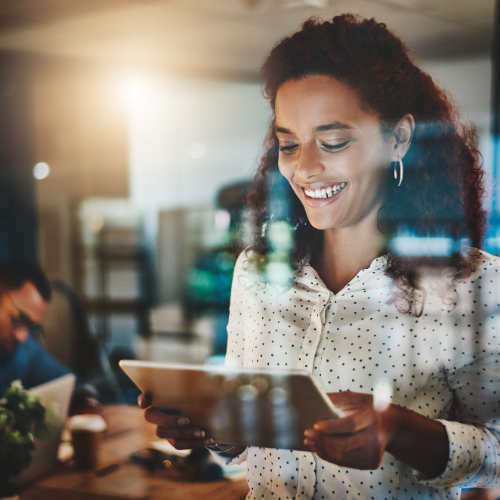 A young professional woman with curly hair, wearing a white polka-dotted blouse, smiling as she looks at a tablet in a modern office setting. The scene is softly lit, with reflections from a glass surface and blurred colleagues working in the background, creating a warm and focused atmosphere.