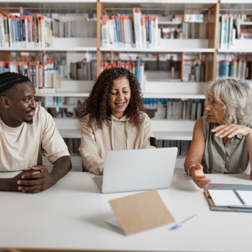 A diverse group of three people sitting at a white table in a library, engaging in a discussion. A middle-aged woman gestures while speaking, a younger woman with curly hair smiles while working on a laptop, and a man listens attentively with his hands folded on the table. The background features shelves filled with books, creating a collaborative and educational atmosphere.